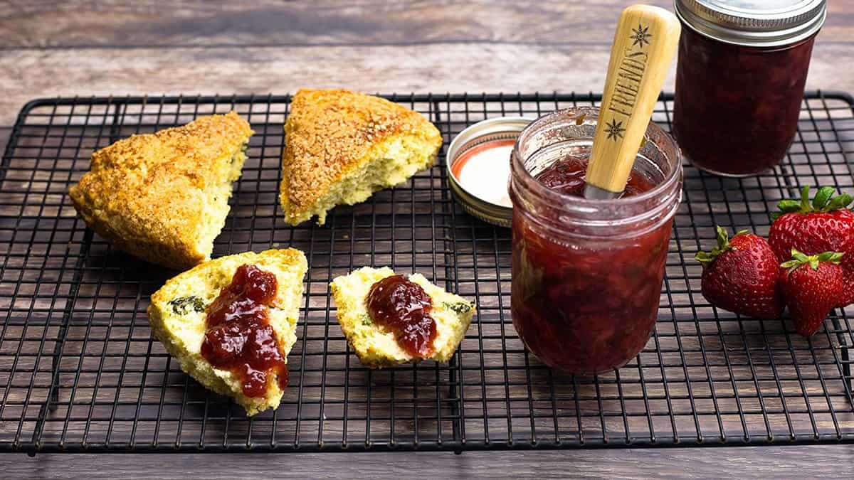 Strawberry Basil Preserves in an open jar with a wooden handled spreader, next to basil scones spread with the preserves, a sealed jar, and fresh strawberries on a cooling rack.