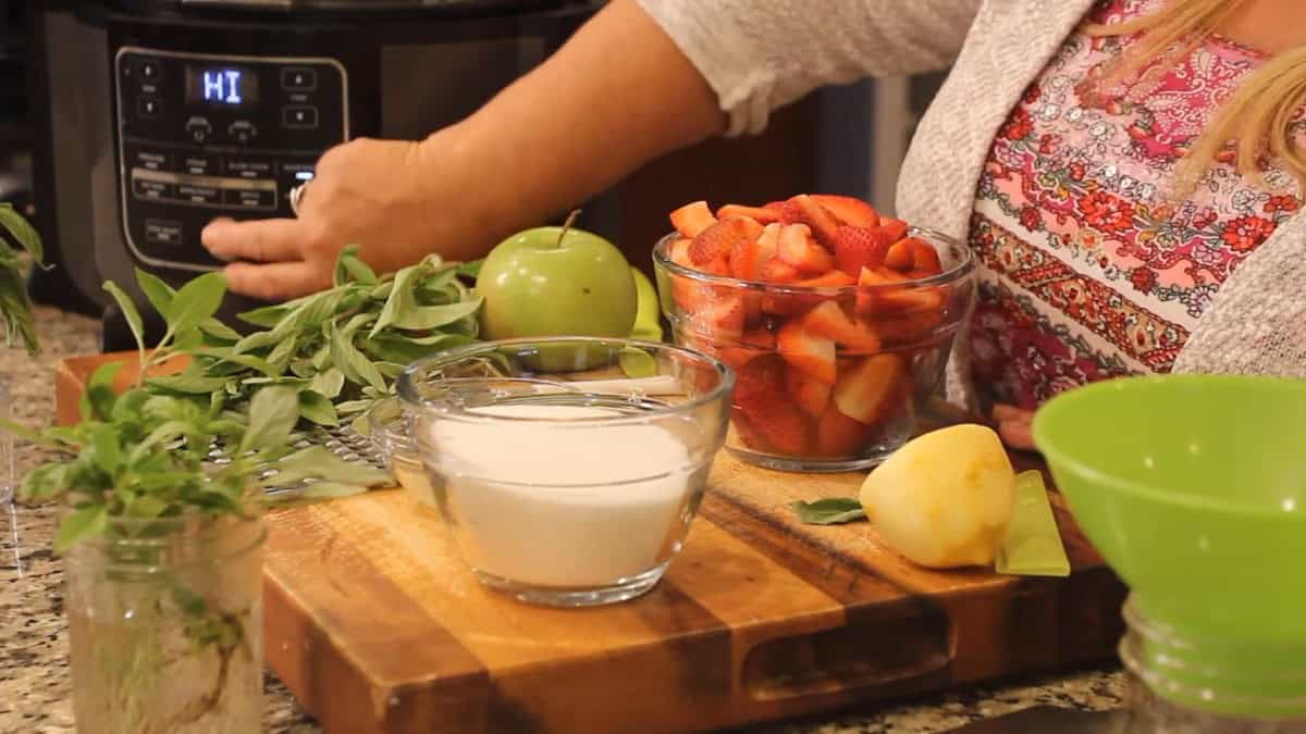 The ingredients for Strawberry Basil Preserves on a wooden board in front of the Ninja Foodi: fresh basil, a Granny Smith apple, a bowl of quartered strawberries, a bowl of sugar, a lemon, and a grater.