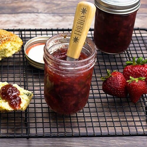 A jar of Strawberry Basil Preserves with a wooden handled spreader resting in it, a second sealed jar, fresh strawberries, and basil flecked scones on a black cooling rack.