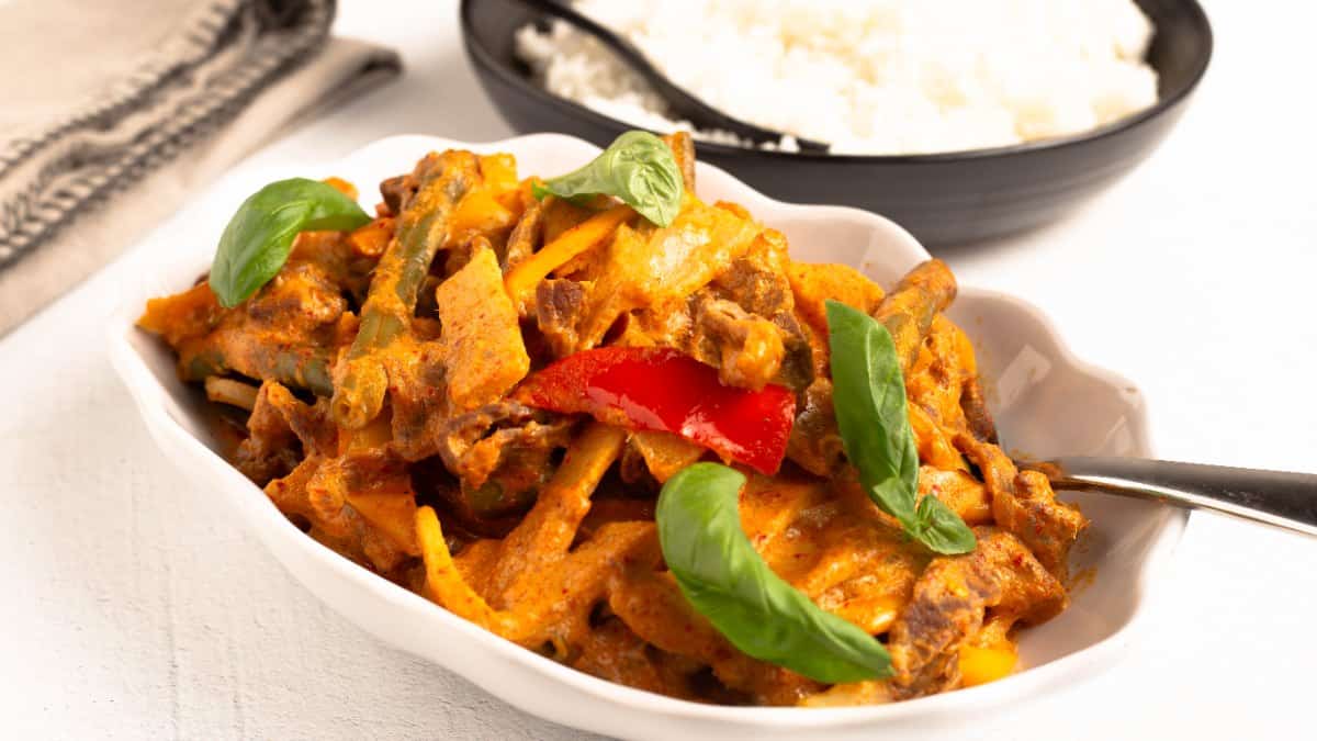 Thai Red Curry Beef in a white scalloped serving dish, with a spoon resting in the creamy red curry sauce, alongside a black bowl of steamed white rice and a striped kitchen towel.