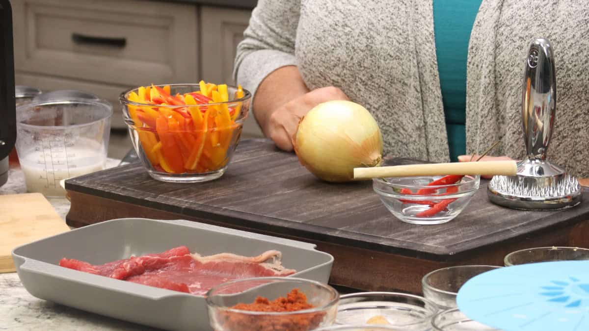 Ingredients for Thai Red Curry Beef arranged on a wooden board, including a glass bowl of orange and yellow mini sweet pepper strips, a whole sweet onion, a stalk of lemongrass, a small bowl of red Thai chilies, a meat mallet, a tray of thinly sliced raw beef, a bowl of red curry paste, and a measuring cup of coconut milk.
