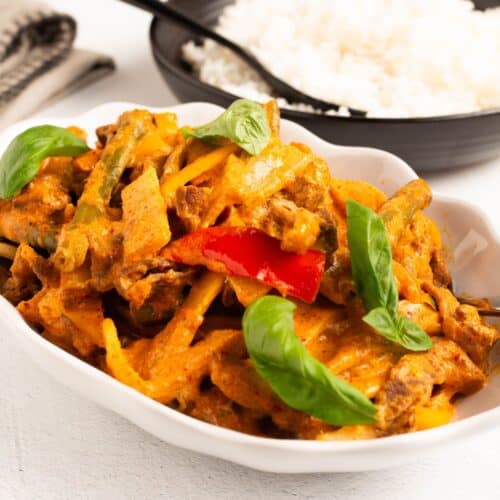 Thai Red Curry Beef served in a white scalloped dish, with strips of beef, pale bamboo shoots, green beans, and red and yellow pepper strips in a creamy red curry sauce, finished with fresh basil leaves. A black bowl of white rice sits behind it.