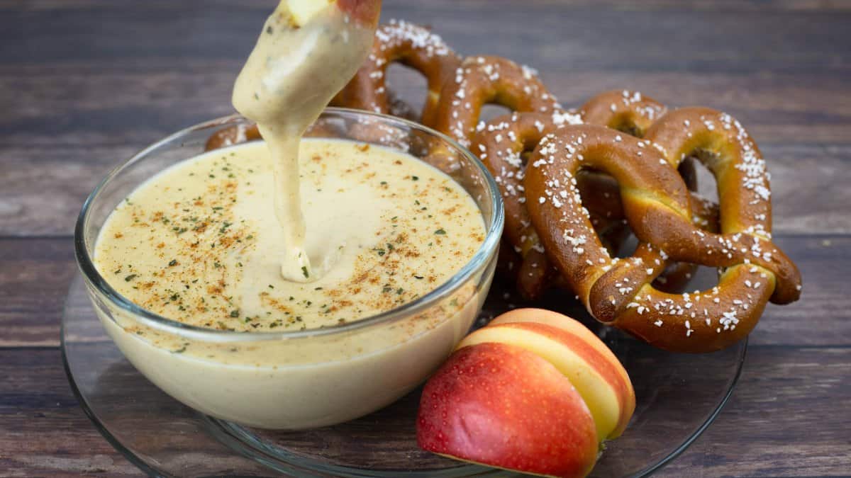Beer Cheese Dip in a glass bowl dusted with spices and parsley, with an apple slice dipping into the smooth cheese sauce beside soft pretzels and a red apple.