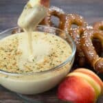 Close up of an apple slice being dipped into a bowl of smooth Beer Cheese Dip, with salted soft pretzels and a red apple on the side.