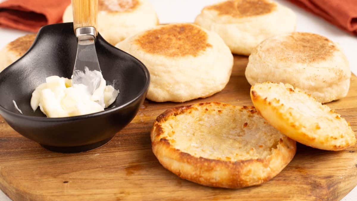 Golden No Knead English Muffins on a round wooden board, one split open and toasted, next to a small black bowl of butter with a spreader.
