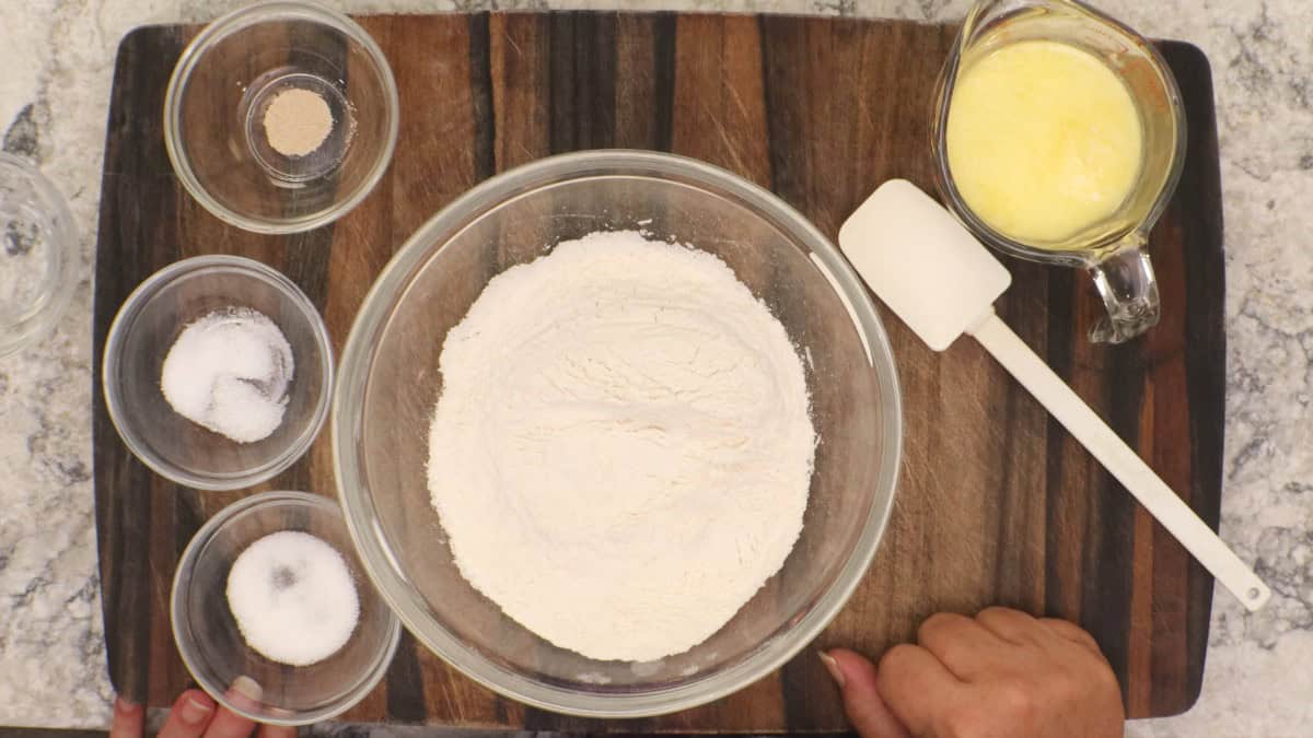 Ingredients for No Knead English Muffins: a glass bowl of bread flour, small bowls of yeast, sugar and salt, a measuring cup of warm milk with melted butter, and a white spatula on a wooden board.