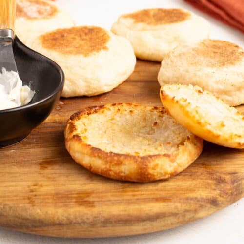 Close-up of toasted, fork-split No Knead English Muffins on a wooden board with more whole muffins and a bowl of butter behind them.