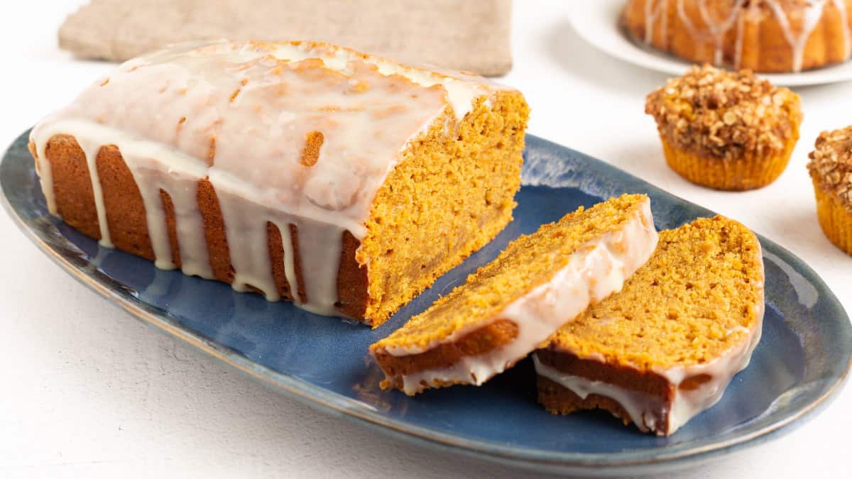 Glazed Pumpkin Bread on a blue oval platter with two slices cut, white chocolate orange glaze dripping down the sides, and pumpkin muffins in the background.