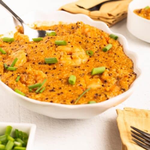 Close view of Shrimp & Grits (Possible cooker Pro in a white dish with a serving spoon, showing shrimp, quinoa and green onions in creamy grits.