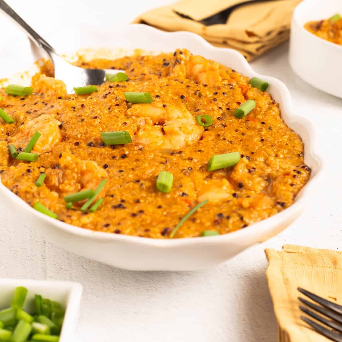 Close view of Shrimp & Grits (Possible cooker Pro in a white dish with a serving spoon, showing shrimp, quinoa and green onions in creamy grits.